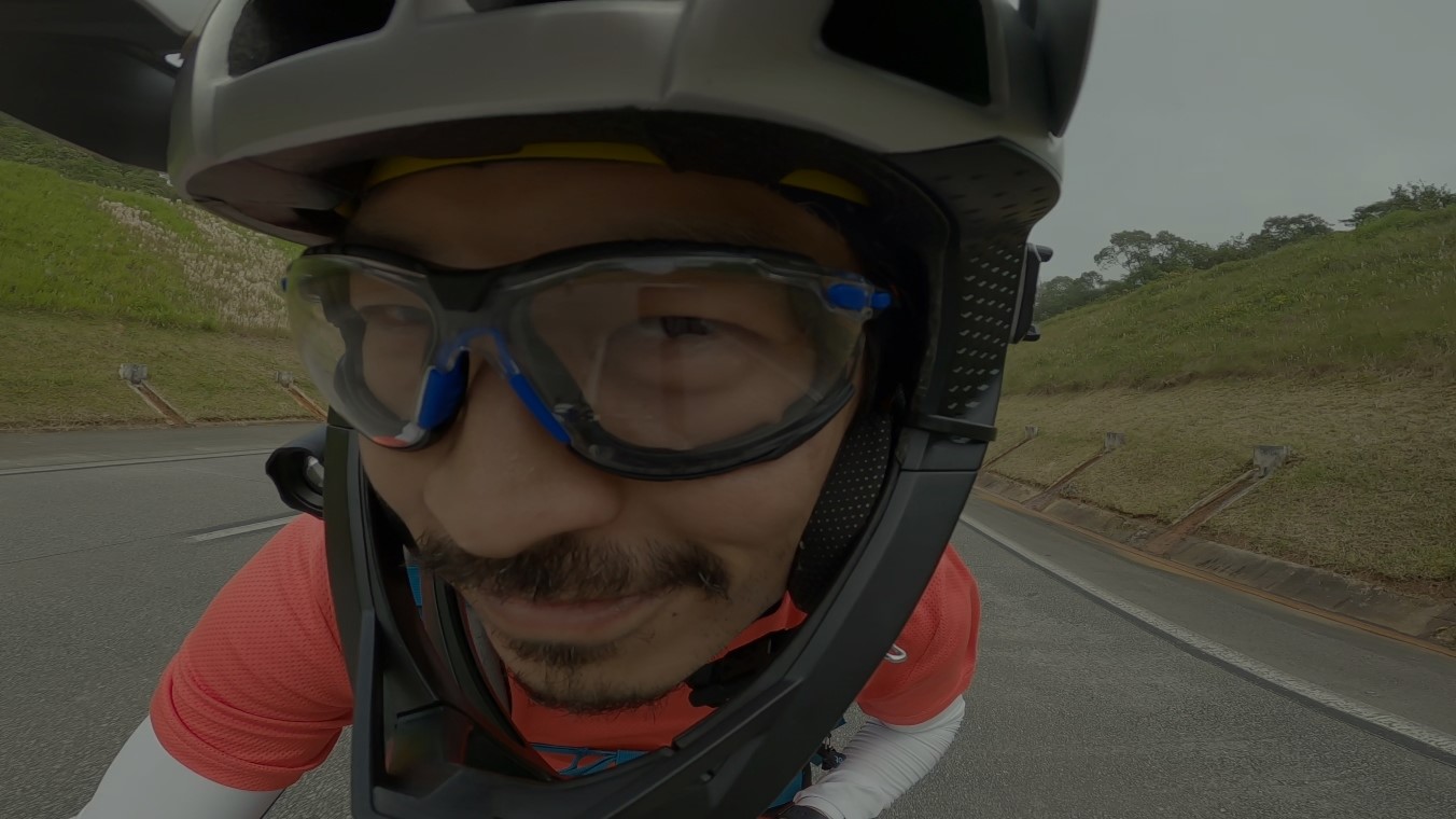 Super close no rosto sorridente do Itiro, que está de capacete fechado e óculos de proteção. No meio de uma estrada asfaltada.